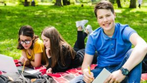 Un grupo de jóvenes estudiantes estudia en el césped de un parque, concentrados en sus tareas mientras disfrutan del entorno natural. Esta escena refleja cómo los Summer Camps en EE.UU. combinan actividades educativas con un ambiente relajado, favoreciendo el aprendizaje y las conexiones.